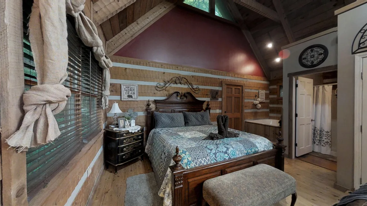 An inviting bedroom scene with a king bed adorned with decorative pillows and an ornate headboard, complemented by a nightstand and lamp. Wood accents and a window with blinds provide a sense of comfort and privacy, offering a glimpse into restful moments.