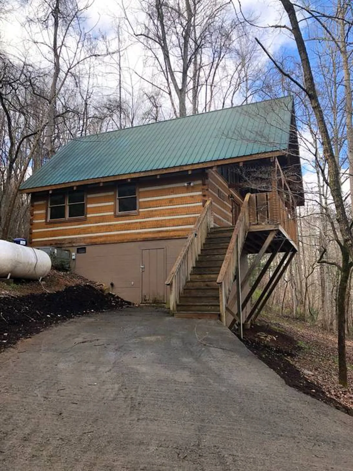 A cozy log cabin structure with a green metal roof and wooden steps, nestled among trees. The setting suggests easy access to the outdoors for hiking or morning coffee and emphasizes the seamless connection between the cabin and its natural surroundings.