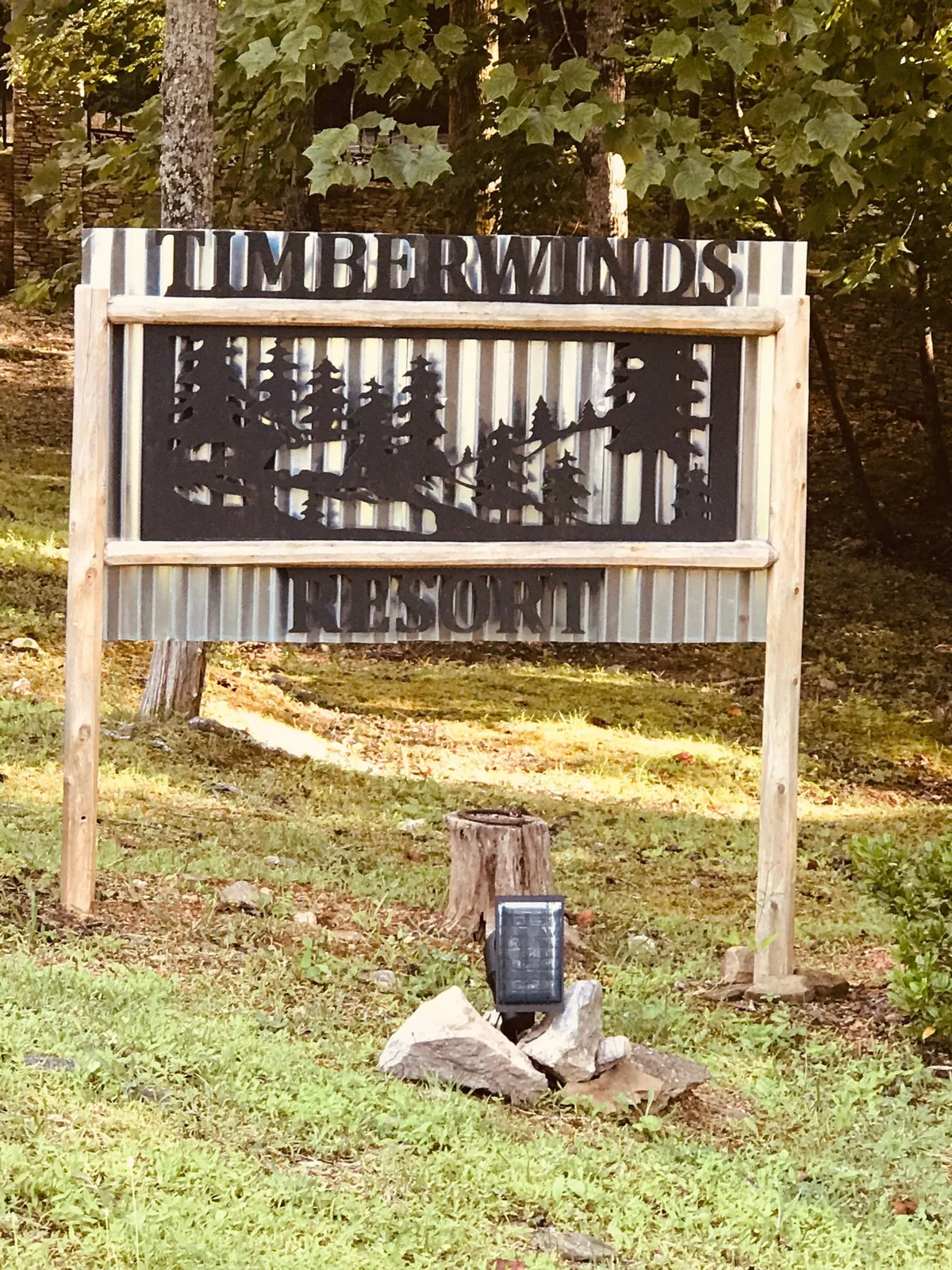 The Timber Winds Resort sign surrounded by towering trees and lush landscape, welcoming guests to an inviting, natural retreat. The wide view hints at walking paths and the nearby natural beauty to explore.