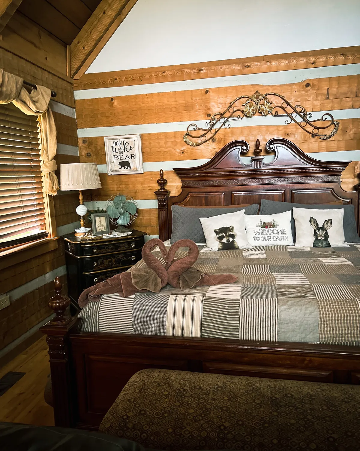 A cozy bedroom scene with a wooden bed frame, decorative pillows, neatly arranged towel art, and inviting wall art. The room exudes warmth, comfort, and attention to detail, with soft lighting enhancing the atmosphere.