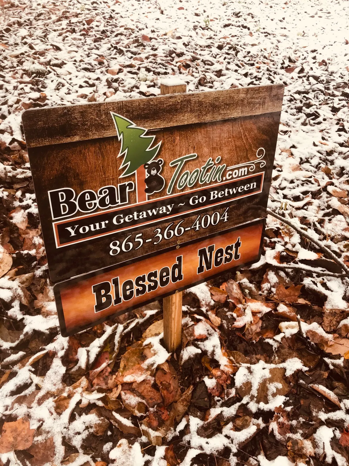 A sign featuring the 'Bear Tootin' logo and 'Blessed Nest' text stands amidst fallen leaves and a light layer of snow, creating a serene and inviting scene. The setting hints at peaceful family gatherings and the welcoming atmosphere of the cabin's location.