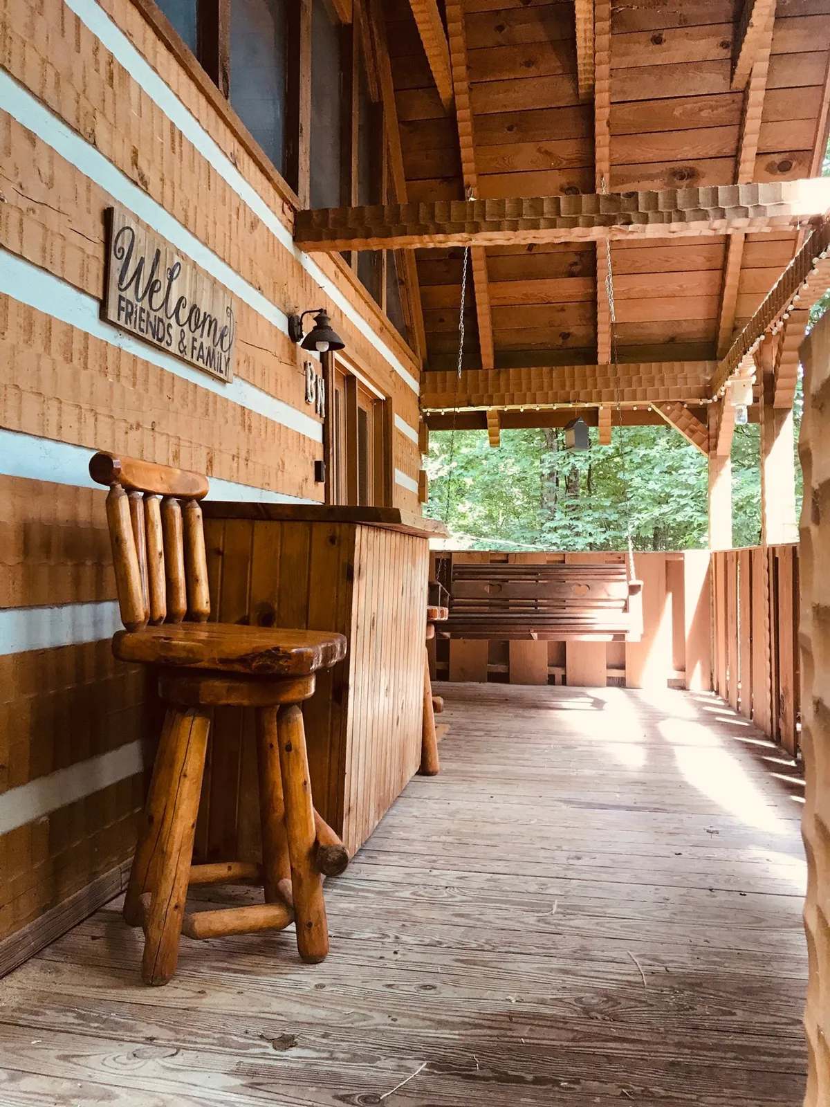 A rustic exterior bar area with a handcrafted stool, welcome sign, and inviting wooden accents. The space is set up for social gatherings, offering a place for friends to relax together while enjoying the natural surroundings and sunsets.