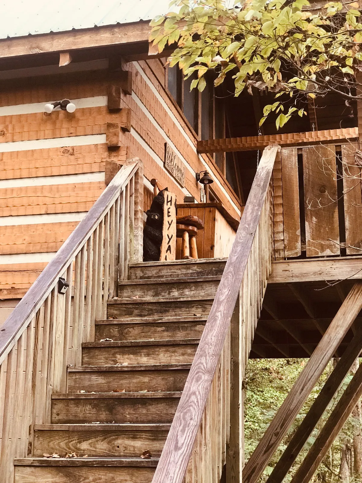 A friendly exterior entryway with a wooden staircase, welcome sign, and a black bear sculpture, inviting guests into the property. The natural wood and bear motif set a welcoming, adventurous tone for arriving friends.